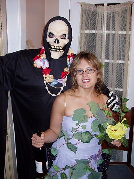 man in skeleton mask with flower lei and a woman wrapped in grape vines with a bottle of wine